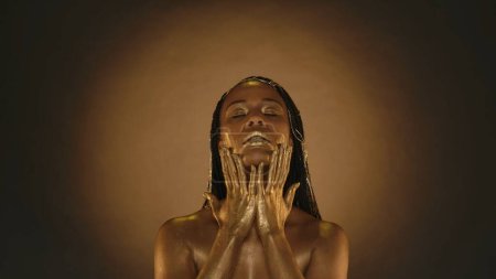 Photo for A woman with creative golden make up and golden colored skin on a brown background with circular light. An African American woman with her eyes closed touches her face with her hands smeared in gold - Royalty Free Image