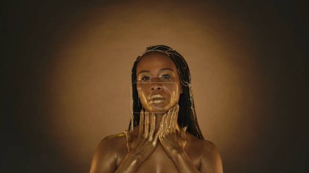 Foto de Una mujer afroamericana se toca el cuello con las manos manchadas de pintura dorada. Una mujer afroamericana con los ojos cerrados toca su cara con las manos manchadas de pintura dorada - Imagen libre de derechos