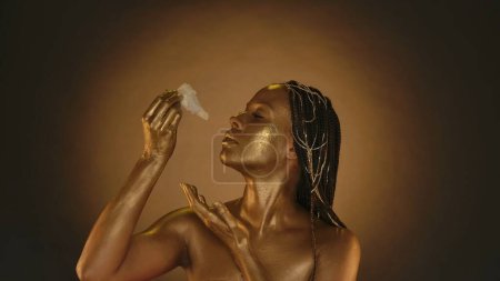 Foto de Una mujer afroamericana con piel dorada trae un capullo de gladiolo a su cara, recogiendo néctar, rocío y humedad vital de ella. Mujer con piel dorada en estudio sobre fondo marrón con - Imagen libre de derechos