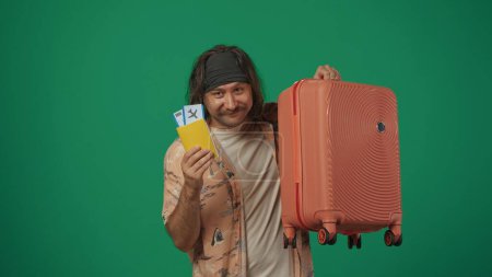 Téléchargez les photos : Concept créatif itinérant. Voyageur homme en tenue décontractée tenant billets d'avion et valise, heureux sourire expression du visage, en regardant la caméra. Isolé sur fond vert. - en image libre de droit
