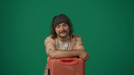 Téléchargez les photos : Concept créatif itinérant. Voyageur homme en vêtements décontractés posant avec valise, souriant en regardant la caméra. Isolé sur fond vert. - en image libre de droit