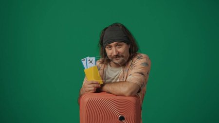 Photo for Travelling creative concept. Man traveller in casual clothing with suitcase holding flight tickets and passport, smiling face expression. Isolated on green background. - Royalty Free Image