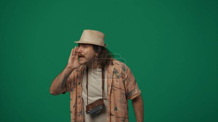 Téléchargez les photos : Concept créatif itinérant. Voyageur homme en vêtements décontractés et chapeau de paille avec une vieille caméra de film regardant le côté et crie. Isolé sur fond vert. - en image libre de droit