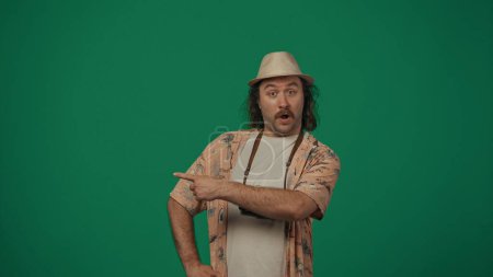 Photo for Travelling creative concept. Man traveller in casual clothing and straw hat with old film camera pointing his hand at the side, surprised face expression. Isolated on green background. - Royalty Free Image