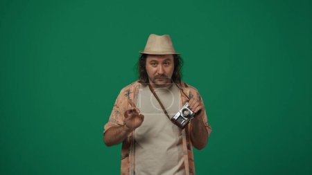 Téléchargez les photos : Concept créatif itinérant. Voyageur homme en vêtements décontractés et chapeau de paille avec vieille caméra de film sur le cou en regardant la caméra avec pose drôle. Isolé sur fond vert. - en image libre de droit