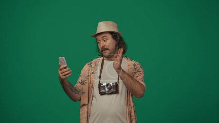 Téléchargez les photos : Concept créatif itinérant. Voyageur homme en vêtements décontractés et chapeau de paille parler sur smartphone par un appel vidéo. Isolé sur fond vert. - en image libre de droit