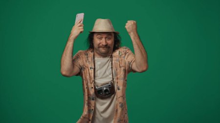 Photo for Travelling creative concept. Man traveller in casual clothing and straw hat holding smartphone, hands up and happy face expression. Isolated on green background. - Royalty Free Image