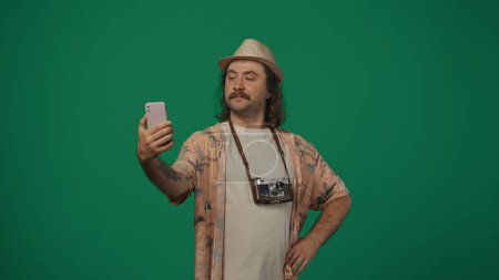 Photo for Travelling creative concept. Man traveller in casual clothing and straw hat with old film camera taking selfie on smartphone, smiling. Isolated on green background. - Royalty Free Image