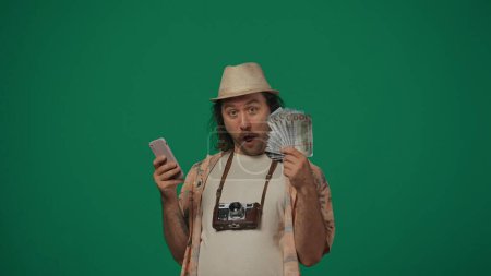 Photo for Travelling creative concept. Man in casual clothing and straw hat with old film camera holding money bills and smartphone, amazed face expression. Isolated on green background. - Royalty Free Image