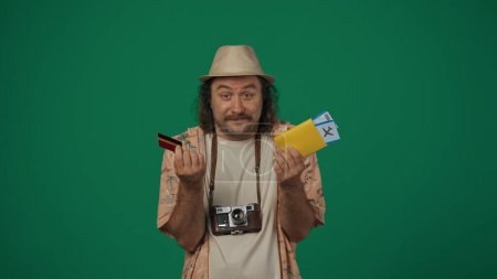 Téléchargez les photos : Concept créatif itinérant. Homme en tenue décontractée et chapeau de paille tenant billets d'avion, passeport et carte de crédit, souriant. Isolé sur fond vert. - en image libre de droit