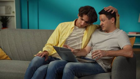 Téléchargez les photos : Fusillade d'un couple homosexuel à la maison. L'un est assis sur le canapé, travaillant sur l'ordinateur portable, expliquant quelque chose à son partenaire qui est assis à côté de lui. Ils s'embrassent. LGBT, égalité, éducation, fierté - en image libre de droit