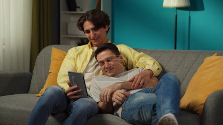 Téléchargez les photos : Fusillade d'un couple homosexuel à la maison. Ils s'allongent sur le canapé, regardent des photos ou des vidéos sur une tablette, s'embrassent, se sourient et s'aiment mutuellement. LGBT, égalité, contenu éducatif, fierté - en image libre de droit