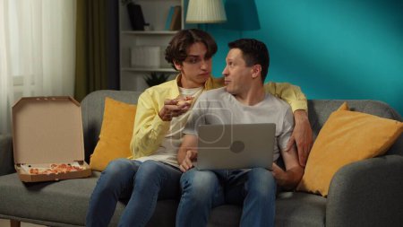 Téléchargez les photos : Fusillade d'un couple homosexuel à la maison. Ils sont assis sur le canapé, regardent quelque chose sur un ordinateur portable, mangent de la pizza, se serrent dans leurs bras et se parlent chaleureusement. LGBT, égalité, contenu éducatif, fierté. - en image libre de droit