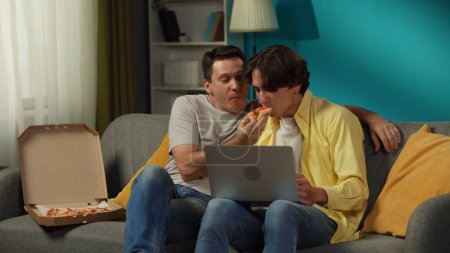 Foto de Un disparo de una pareja homosexual en casa. Están sentados en el sofá, mirando algo en un portátil, comiendo pizza, abrazándose y hablando calurosamente entre sí. LGBT, igualdad, contenido educativo, orgullo. - Imagen libre de derechos