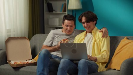 Foto de Un disparo de una pareja homosexual en casa. Están sentados en el sofá, mirando algo en un portátil, comiendo pizza, abrazándose y hablando calurosamente entre sí. LGBT, igualdad, contenido educativo, orgullo. - Imagen libre de derechos