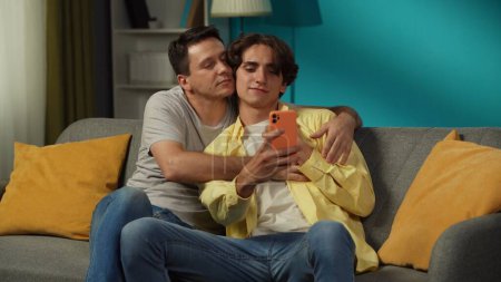 Photo for Shot of two homosexual men, couple at home. They are sitting on the couch, hugging, taking selfies, expressing love and affection to each other. LGBT, equality, educational content, pride. - Royalty Free Image