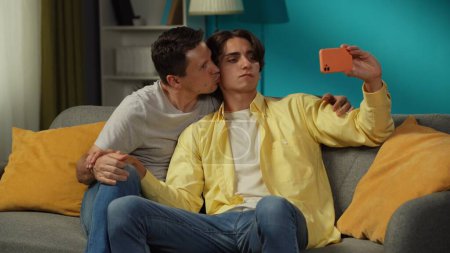 Foto de Un disparo de dos hombres homosexuales, una pareja en casa. Están sentados en el sofá, abrazándose, tomando selfies, expresándose amor y afecto el uno al otro. LGBT, igualdad, contenido educativo, orgullo. - Imagen libre de derechos