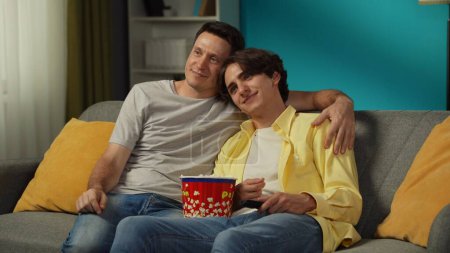 Foto de Un disparo de una pareja homosexual en casa. Están sentados en el sofá, viendo la televisión juntos, comiendo palomitas de maíz, abrazándose, sonriendo y expresándose amor el uno al otro. LGBT, igualdad, contenido educativo, orgullo - Imagen libre de derechos