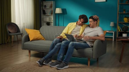 Foto de Una foto grande de una pareja homosexual en casa. Uno está sentado en el sofá, trabajando en el portátil, explicando algo a su pareja que se sienta a su lado. Se están abrazando. LGBT, igualdad, orgullo. - Imagen libre de derechos