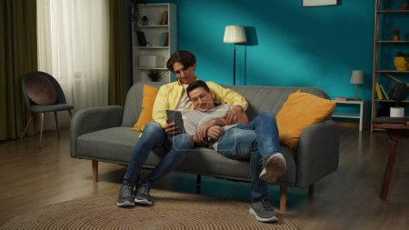 Téléchargez les photos : Plan complet d'un couple homosexuel à la maison. Ils s'allongent sur le canapé, regardent des photos ou des vidéos sur une tablette, s'embrassent, se sourient et s'aiment mutuellement. LGBT, éducatif, fier. - en image libre de droit