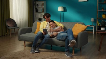 Photo for Full-size shot of a homosexual couple at home. They are laying on the couch, watching photos or videos on a tablet, hugging, smiling and expressing love to each other. LGBT, educational, pride. - Royalty Free Image