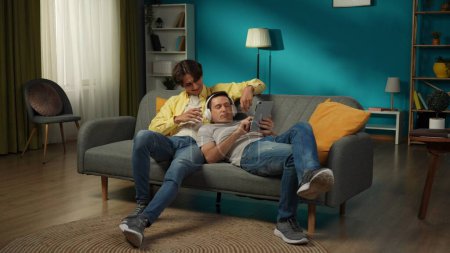 Téléchargez les photos : Plan complet d'un couple homosexuel à la maison. Ils sont allongés sur le canapé, regardant des photos ou des vidéos sur une tablette. L'un partage leurs écouteurs. LGBT, égalité, contenu éducatif, fierté. - en image libre de droit