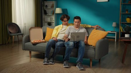 Photo for Full-size shot of a homosexual couple at home. They are sitting on the couch, watching something on a laptop, eating pizza, hugging and talking warmly to each other. LGBT, equality, educational, pride - Royalty Free Image