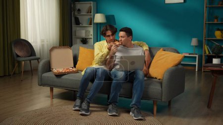 Foto de Una foto grande de una pareja homosexual en casa. Están sentados en el sofá, mirando algo en un portátil, comiendo pizza, abrazándose y hablando calurosamente entre sí. LGBT, igualdad, educación, orgullo - Imagen libre de derechos
