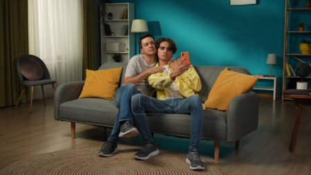 Photo for Full-size shot of two homosexual men, couple at home. They are sitting on the couch, hugging, taking selfies, expressing love and affection to each other. LGBT, equality, educational content, pride. - Royalty Free Image