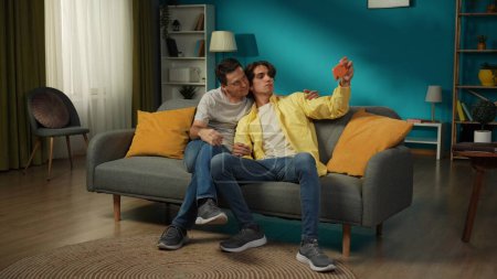 Photo for Full-size shot of two homosexual men, couple at home. They are sitting on the couch, hugging, taking selfies, expressing love and affection to each other. LGBT, equality, educational content, pride. - Royalty Free Image