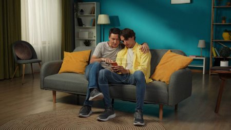 Photo for Full-size shot of a homosexual couple at home. They are laying on the couch, watching photos or videos on a smartphone, hugging, smiling and expressing love to each other. LGBT, educational, pride. - Royalty Free Image
