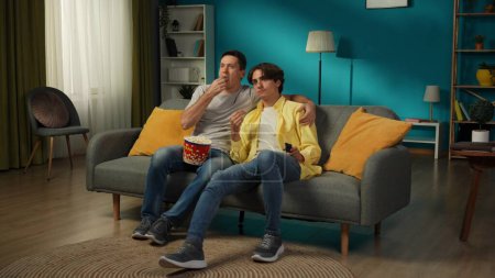 Foto de Una foto grande de una pareja homosexual en casa. Están sentados en el sofá, viendo la televisión juntos, comiendo palomitas de maíz, abrazándose, sonriendo y expresándose amor el uno al otro. LGBT, educación, orgullo. - Imagen libre de derechos