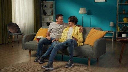 Foto de Una foto grande de una pareja homosexual en casa. Están sentados en el sofá, viendo la televisión juntos, comiendo palomitas de maíz, abrazándose, sonriendo y expresándose amor el uno al otro. LGBT, educación, orgullo. - Imagen libre de derechos