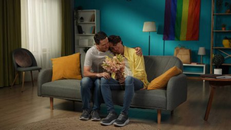 Photo for Full-size shot of a homosexual couple at home. One has just gave flowers, bouquet to their partner, expressing love and affection. The parnters are hugging tenderly. LGBT, equality, educational, pride - Royalty Free Image