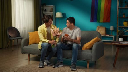 Photo for Full-size shot of a homosexual couple at home. One is giving flowers, bouquet to their partner, saying compliments, wishes, warm words, expressing love and affection. LGBT, equality, educational - Royalty Free Image