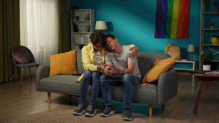 Foto de Foto de tamaño completo de una pareja homosexual sentada en el sofá juntos, abrazándose, consolándose, tomándose de la mano, expresando amor y afecto. LGBT, igualdad, contenido educativo, orgullo. - Imagen libre de derechos