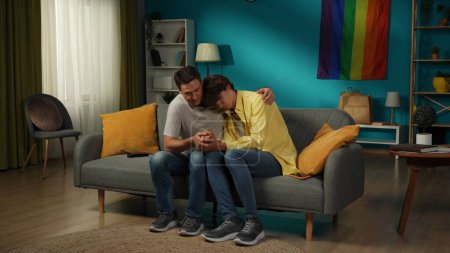 Photo for Full-size shot of a homosexual, pansexual, bisexual male couple sitting on the couch, hugging, holding hands. One is comforting their partner. LGBT, equality, educational content, pride. - Royalty Free Image