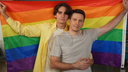 Foto de Mediana toma de una pareja homosexual, bisexual, pansexual, transgénero mirando a la cámara, abrazando y sosteniendo la bandera LGBT para fomentar el amor, la igualdad, la libertad. Contenido educativo, orgullo, valentía. - Imagen libre de derechos