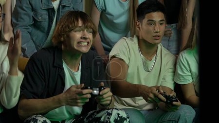 Group of teens, young people, friends sitting and standing around the TV and cheering, encouraging two guys, boys who are playing a console, gaming, play-fighting. Gamer, teenage, youth content.