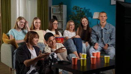 Foto de Grupo de adolescentes, jóvenes, amigos sentados en un sofá, de fiesta, viendo televisión, cine, espectáculo. Están comiendo palomitas de maíz, riendo y charlando, bromeando. Publicidad, adolescentes, contenido juvenil. - Imagen libre de derechos