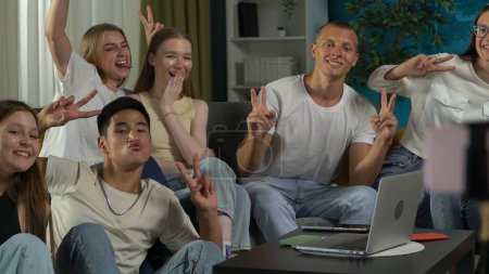 Photo for Medium shot of a group of teens, young people, friends sitting around the table, using a smartphone on a tripod to take a picture, photo together, showing peace gesture. Advertisement, gen z, social. - Royalty Free Image