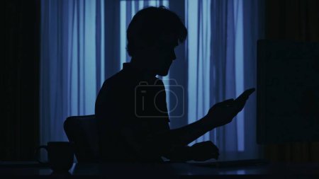 Photo for Portrait of male silhouette in the dark apartment. Everyday life creative concept. Man at the desk in the dark room working, reading something on his smartphone. - Royalty Free Image