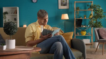 Foto de El hombre lee un libro mientras está sentado en casa en el sofá. El concepto de felicidad familiar, ocio, descanso en casa - Imagen libre de derechos