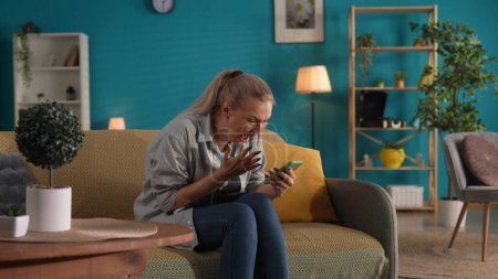 Photo for A woman holds a phone and screams with joy. A woman won an online game or received a prize. Portrait of a happy, shocked woman with a phone in her hands on the sofa in the living room - Royalty Free Image