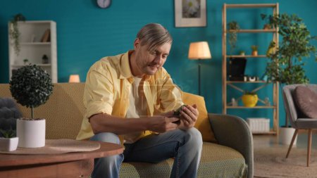 Foto de Un hombre que usa un teléfono inteligente mientras está sentado en el sofá en casa de cerca. Un hombre se desplaza a través de su alimentación en las redes sociales, mira fotos, videos y juega un juego en línea. Concepto de felicidad familiar - Imagen libre de derechos