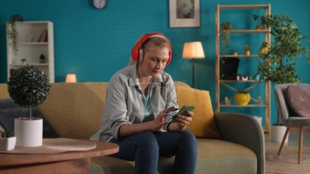 Foto de Una mujer que usa auriculares inalámbricos rojos y sostiene un teléfono está sentada en el sofá de su casa. - Imagen libre de derechos
