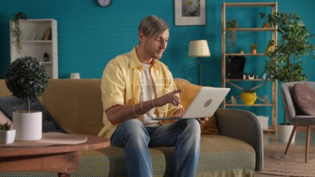 Foto de Un hombre con ropa casual se sienta en un sofá en la sala de estar con una computadora portátil en su regazo. Un hombre trabaja, escribe un texto, un mensaje, chats, juega juegos en línea. Trabajo remoto, freelancer - Imagen libre de derechos