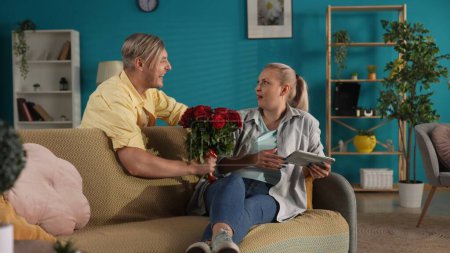Téléchargez les photos : Je fais la surprise. Un homme présente un bouquet de roses rouges à une femme assise sur un canapé avec une tablette. Anniversaire, Fête des femmes, Saint Valentin - en image libre de droit