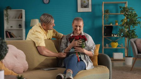 Téléchargez les photos : Je fais la surprise. Un homme présente un bouquet de roses rouges à une femme assise sur un canapé avec une tablette. Anniversaire, Fête des femmes, Saint Valentin - en image libre de droit