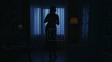 Foto de Retrato de silueta femenina en el apartamento oscuro. Mujer vestida con tacones altos mirándose en el espejo en la sala de estar. Concepto creativo de la vida cotidiana. - Imagen libre de derechos